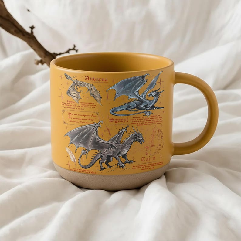 May include: Mustard yellow ceramic mug featuring illustrations of dragons with wings. The mug has a tan base and a curved handle. The mug has text that reads "Anoarna", "Sgaeyl", and "Tatu".