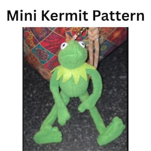 May include: A plush green frog keyring featuring the character Kermit with a pointed collar, long limbs, and large white eyes. The text 'Mini Kermit Pattern' is displayed above the plush toy, which is attached to a metal ring.