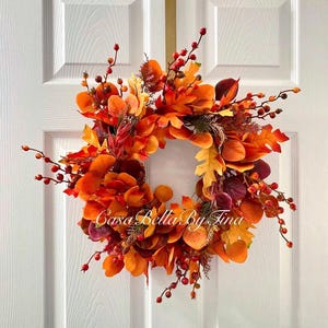 May include: A circular autumn wreath featuring a dense arrangement of faux leaves in shades of orange, yellow, red, and deep burgundy. The wreath is accented with small red berries on thin stems and includes the text Casa Bella By Tina in white script.