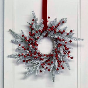 May include: A circular wreath featuring frosted, snow-dusted evergreen branches interspersed with clusters of bright red berries. The wreath is suspended by a vertical dark red velvet ribbon, creating a contrast against the white foliage.