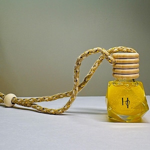 Puede incluir: Una pequeña botella de vidrio amarillo facetado con una tapa de madera y un cordón trenzado. La botella contiene un líquido brillante y tiene un símbolo impreso. El cordón es de color beige y dorado, con una cuenta de madera.