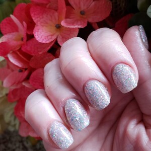 May include: Close-up of a hand with manicured nails. The nails are painted with a sparkly, iridescent polish that reflects light. The hand is positioned in front of a backdrop of pink and red flowers.