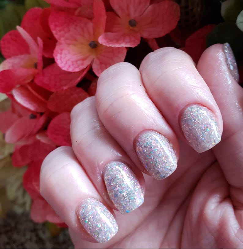 May include: Close-up of a hand with manicured nails. The nails are painted with a sparkly, iridescent polish that reflects light. The hand is positioned in front of a backdrop of pink and red flowers.