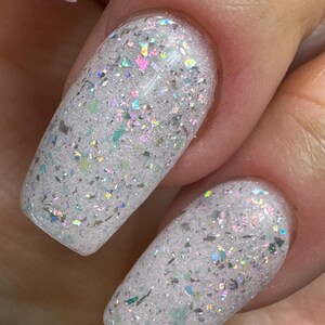 May include: Close-up of two fingernails with white nail polish and iridescent glitter. The glitter reflects multiple colours, including pink, green, and silver. The nails are oval-shaped and have a glossy finish.