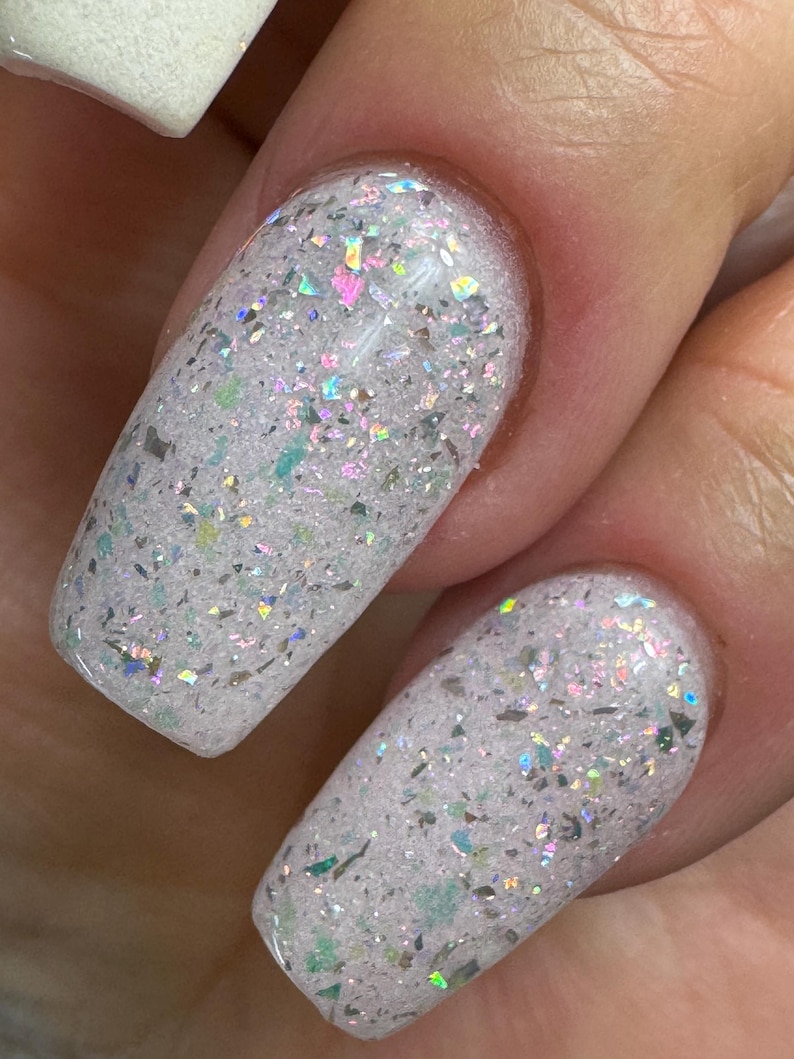 May include: Close-up of two fingernails with white nail polish and iridescent glitter. The glitter reflects multiple colours, including pink, green, and silver. The nails are oval-shaped and have a glossy finish.