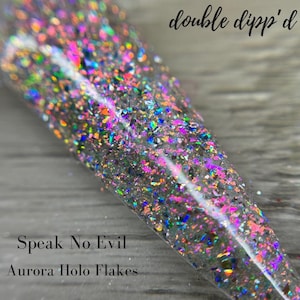 May include: Close-up of a clear container filled with iridescent glitter flakes. The flakes are a mix of colours, including pink, purple, green, and blue. Text on the image reads "double dipp'd", "Speak No Evil", and "Aurora Holo Flakes".