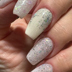 May include: Close-up of a hand with long, square acrylic nails. The nails are painted with a white and iridescent glitter design, creating a sparkling effect. The glitter catches the light, adding a touch of glamour.