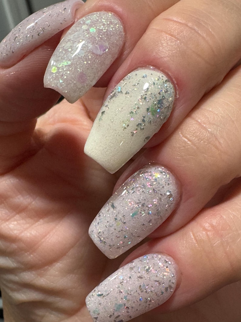 May include: Close-up of a hand with long, square acrylic nails. The nails are painted with a white and iridescent glitter design, creating a sparkling effect. The glitter catches the light, adding a touch of glamour.