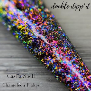 May include: Close-up of a nail with iridescent chameleon flakes. The nail is covered in a mix of colors, including blue, purple, pink, and yellow. The text "double dipp'd" is at the top, and "Cast a Spell Chameleon Flakes" is at the bottom.