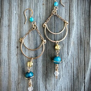 May include: A pair of gold-toned earrings with turquoise and clear crystal accents. Each earring features a crescent-shaped design, a turquoise bead, and a dangling crystal. The earrings are displayed against a weathered wood background.