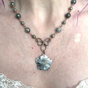 May include: A copper-toned necklace featuring a flower pendant with a pearlescent finish. The necklace has a beaded chain with green and gray beads. The pendant is a five-petal flower design.
