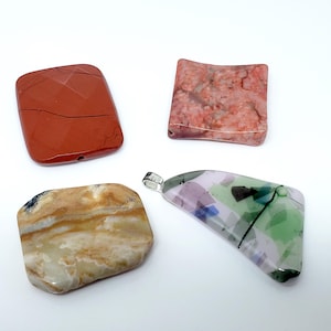 May include: Four colorful pendants and stones on a white background. The pendants include a red-brown rectangular stone, a pink and red square stone, a beige and brown rectangular stone, and a multi-colored glass pendant with a silver bail.