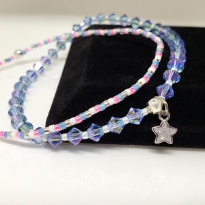 May include: A delicate necklace featuring iridescent blue and clear crystal beads, interspersed with pink and white beads. A silver star charm dangles from the necklace. The necklace is displayed on a black surface.