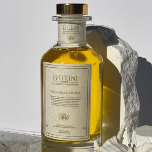 May include: A clear glass bottle filled with yellow liquid, featuring a gold cap and label. The label reads "FATEINI" and "VOIDOKILIA EUPHORIA". The bottle is a home fragrance product, designed in California.