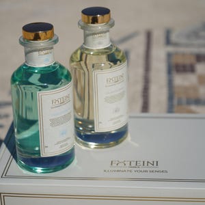 May include: Two glass bottles with gold-colored caps and labels. One bottle contains a teal liquid, the other a clear liquid. The labels read "FXTEINI GREECE ILLUMINATE YOUR SENSES". The bottles sit on a white box with gold trim.