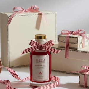 Op de afbeelding: Een heldere glazen fles gevuld met een rode vloeistof, afgedekt met een gouden dop en een rood-wit gestreepte strik. De fles heeft een etiket met tekst. Cadeaudozen met bijpassende strikken en linten staan op de achtergrond.