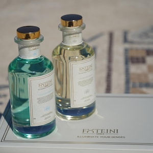 Op de afbeelding: Twee glazen parfumflessen met gouden en zwarte doppen. Eén fles bevat een turquoise vloeistof, de andere een gele vloeistof. Elke fles heeft een wit etiket met de tekst "FXTEINI ILLUMINATE YOUR SENSES". De flessen staan op een wit oppervlak.