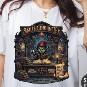 May include: White t-shirt featuring a detailed illustration of a goblin reading a book and holding a glass of wine. The design includes the text "SMUT GOBLIN TEE", "DARK ROMANCE", "READERS CLUB", and "SPICY BOOK LOVER".