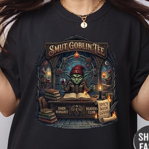 Może przedstawiać: Czarny t-shirt z grafiką przedstawiającą zielonego goblina czytającego książkę, trzymającego kieliszek wina, oraz napisem "SMUT GOBLIN TEE". Dodatkowy tekst zawiera "DARK ROMANCE", "READERS CLUB" i "SPICY BOOK LOVER".