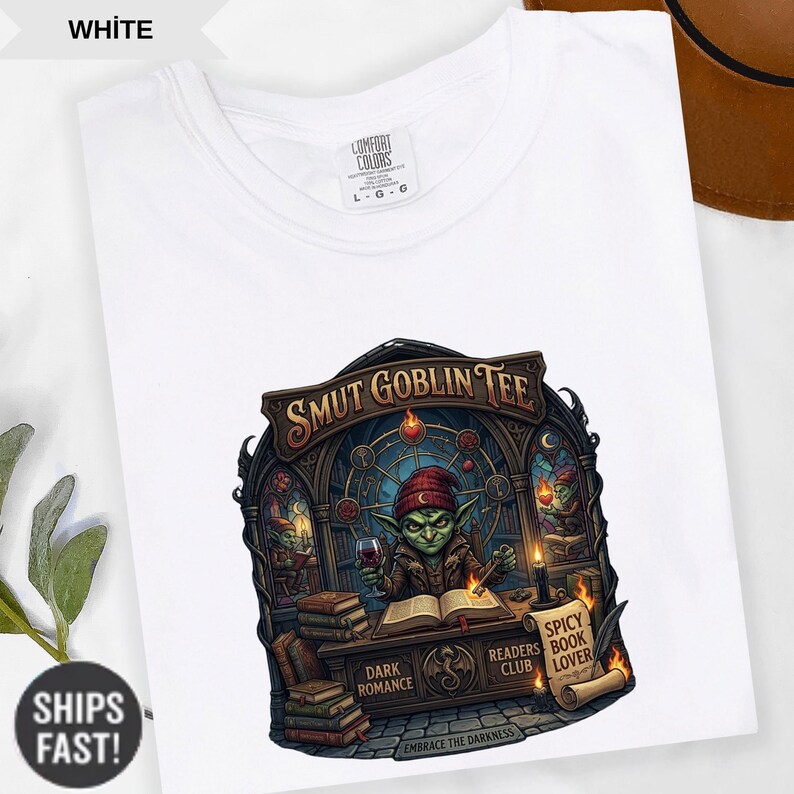 Może przedstawiać: Biała koszulka z grafiką przedstawiającą goblina czytającego książkę, świecę i napis "SMUT GOBLIN TEE". Projekt zawiera frazy "Dark Romance", "Readers Club" i "Spicy Book Lover".