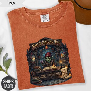 Może przedstawiać: Pomarańczowa koszulka Comfort Colors z grafiką z napisem "SMUT GOBLIN TEE". Grafika przedstawia zielonego goblina trzymającego kieliszek wina, z książkami i świecami. Koszulka zawiera r&oacute;wnież tekst "DARK ROMANCE" i "SPICY BOOK LOVER".