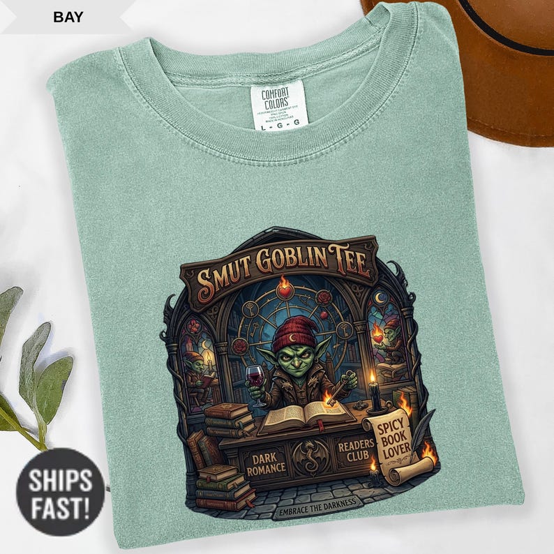 Może przedstawiać: Jasnozielona koszulka Comfort Colors z grafiką. Grafika zawiera tekst "SMUT GOBLIN TEE" nad ilustracją goblina czytającego książkę i trzymającego kieliszek wina. Dodatkowy tekst zawiera "DARK ROMANCE", "READERS CLUB" i "SPICY BOOK LOVER".