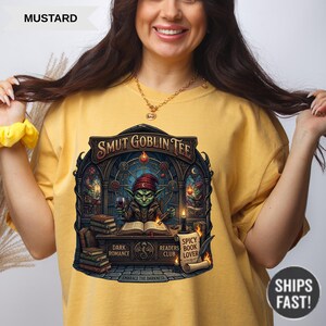 Może przedstawiać: Musztardowa koszulka z napisem "SMUT GOBLIN TEE" nad grafiką goblina czytającego książkę. Grafika zawiera słowa "DARK ROMANCE", "READERS BOOK CLUB" i "SPICY BOOK LOVER". Koszulka ma r&oacute;wnież napis "EMBRACE THE DARKNESS".