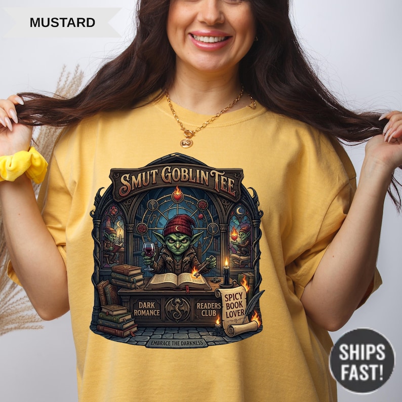Może przedstawiać: Musztardowa koszulka z napisem "SMUT GOBLIN TEE" nad grafiką goblina czytającego książkę. Grafika zawiera słowa "DARK ROMANCE", "READERS BOOK CLUB" i "SPICY BOOK LOVER". Koszulka ma r&oacute;wnież napis "EMBRACE THE DARKNESS".
