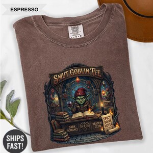 Może przedstawiać: Koszulka w kolorze espresso z grafiką z napisem "SMUT GOBLIN TEE". Projekt przedstawia zielonego goblina trzymającego kieliszek wina, z książkami i świecami. Koszulka wykonana jest z materiału Comfort Colors.