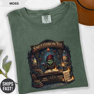 Może przedstawiać: Koszulka Comfort Colors w kolorze mchu z grafiką czytającego goblina i napisem "SMUT GOBLIN TEE". Projekt zawiera słowa "Dark Romance", "Readers Club" i "Spicy Book Lover".