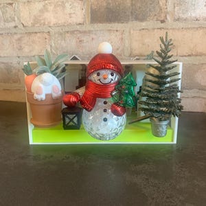 May include: A festive holiday display featuring a snowman with a red hat and scarf, holding a green Christmas tree. A bunny in a pot with carrots and a small Christmas tree are also included. The display is set on a green base.