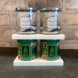 May include: Two-tiered white candle holder with four scented candles. The top level holds two Marshmallow Fireside candles, 8 oz each. The bottom level holds two Fresh Balsam candles. The candles have silver lids and decorative labels.