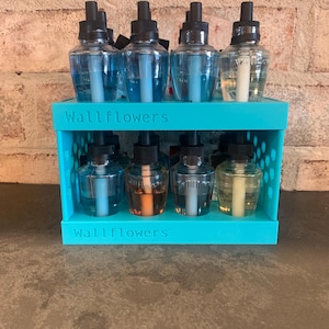 May include: A teal-colored organizer holds eight glass Wallflowers fragrance refills. The refills contain various colored liquids, including blue, amber, and clear. The text "Wallflowers" is printed on the front of the organizer.