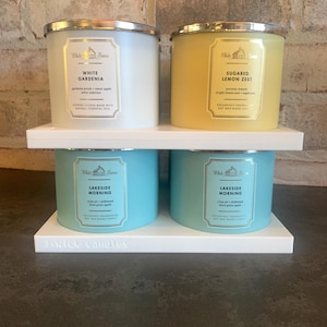 May include: Four scented 3-wick candles on a white shelf. Two candles are white with silver lids, labeled "White Gardenia" and "Sugared Lemon Zest." The other two are blue, labeled "Lakeside Morning." The shelf reads "3-Wick Candles."
