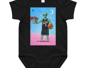 May include: Black baby bodysuit featuring a colorful graphic of an alien playing basketball. The alien wears a jersey with "San Antonio" and the number "1". The image includes a basketball hoop, a crescent moon, and the text "EL WEMBALIEN".