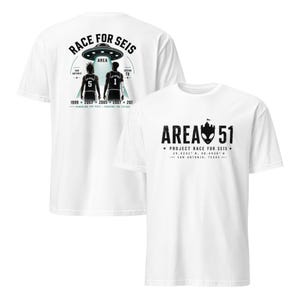 Pode incluir: Duas camisetas brancas com gráficos pretos. Uma camiseta apresenta um design da Área 51 com coordenadas e texto. A outra camiseta tem um design de OVNI com duas figuras e o texto "RACE FOR SEIS".