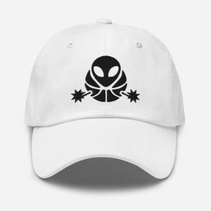 Puede incluir: Gorra de béisbol blanca con un gráfico negro de una cabeza alienígena sobre una pelota de baloncesto con espuelas en forma de estrella. La gorra tiene una visera curva y un cierre de tela en la parte trasera.