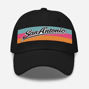 Pode incluir: Boné de beisebol preto com o texto "San Antonio" em itálico, acima de um design de listras horizontais em verde-azulado, rosa e laranja. Uma pequena estrela está no lado esquerdo do texto.