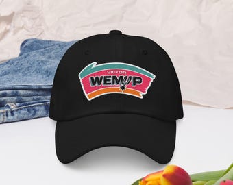 May include: Black baseball cap with the text "VICTOR WEMUP" in pink, white, and teal, with an orange border. The cap has a curved brim and is made of a soft material. The cap is a sports-themed accessory.