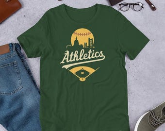 May include: Dark green t-shirt featuring a baseball design with the word "Athletics" in a vintage font. The design includes a baseball, a baseball field, and a city skyline in yellow and white.
