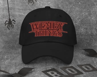 May include: Black baseball cap with the text "WEMBY THINGS" in red, stylized lettering. The cap is a classic six-panel design with a curved brim. The background includes Halloween-themed decorations.
