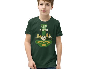 May include: A dark green t-shirt featuring a baseball field design with the text "LIGHT THE GREEN". The design includes the Oakland Athletics logo and city skyline in yellow and white. The shirt is worn by a person.