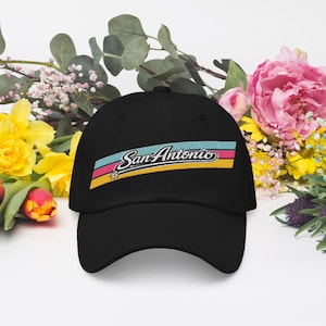 May include: Black baseball cap with the embroidered text "San Antonio" in a colorful design. The text is in white script with a light blue, pink, and yellow stripe design. The cap is surrounded by flowers.