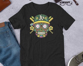 May include: Dark gray t-shirt featuring a colorful sugar skull design with baseball elements. The skull wears a green baseball cap with "A's" in yellow, crossed bats, and baseballs for eyes. Floral accents complete the design.