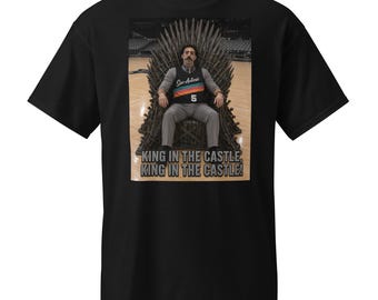 May include: Black t-shirt featuring a graphic of a person seated on a throne. The person wears a jersey with the text "San Antonio" and the number "5". The text "KING IN THE CASTLE, KING IN THE CASTLE!" is printed below the image.
