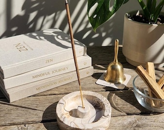 May include: A round, beige and brown marbled incense holder with a central pillar, holding a burning incense stick with visible smoke. Nearby are three stacked books labeled Zen, Mindful Journey, and Meditation Journey, a brass bell, and a glass bowl.