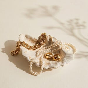 May include: A white and brown marbled shell dish containing a gold-toned chunky chain bracelet, a long strand of white pearls, and two small gold-toned rings with a ribbed texture. The jewelry is arranged inside the decorative shell bowl.