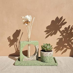 May include: A light green concrete set featuring a rectangular tray, a square vase with a circular cutout holding a glass tube with a white decorative flower, and a small cylindrical planter containing a green succulent plant.