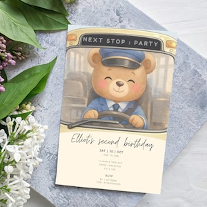 Puede incluir: Una tarjeta de invitación beige claro con un oso de dibujos animados conduciendo un autobús, con el texto "NEXT STOP: PARTY". La tarjeta dice "Elliot's second birthday" con fecha, hora y detalles de RSVP. El oso lleva un uniforme azul.