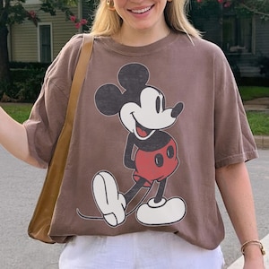 Peut inclure: Un t-shirt marron avec un dessin de Mickey Mouse en noir, rouge et blanc. Le t-shirt est port&eacute; avec un short blanc et une casquette de baseball beige. Le mot "ESPRESSO" est dans un cercle en bas &agrave; gauche.